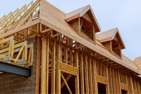 Wooden frame of a house under construction, showing the structure of walls, roof, and dormers against a cloudy sky.